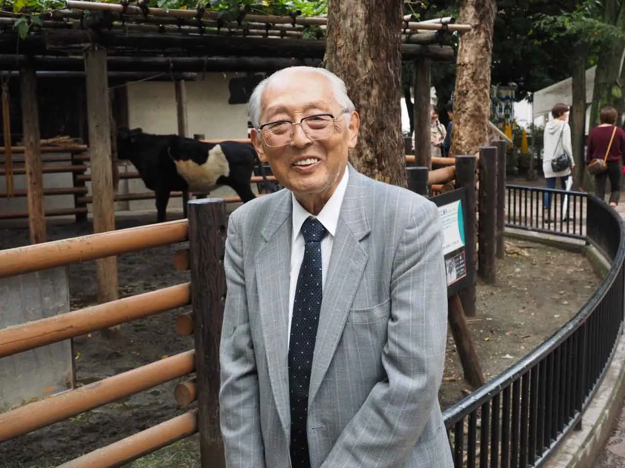 上野動物園の柵の前で笑顔で立つ正田陽一先生。奥の放飼場にウシがいる